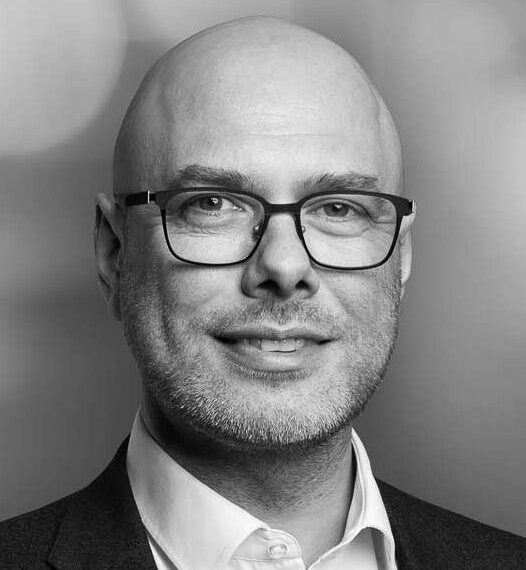 En man med glasögon och kostym ler mot kameran i svartvitt.