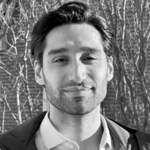 En man står utomhus framför en tegelvägg och trädgrenar, fotograferad i svartvitt.