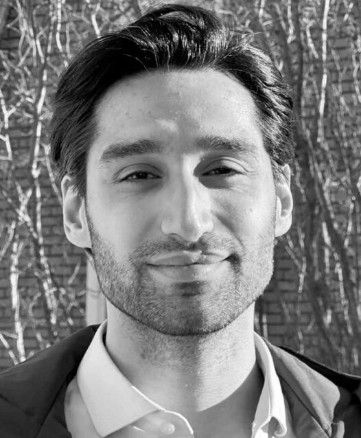 En svartvit porträttbild av en person som ler lätt. Bakgrunden visar en tegelvägg och trädgrenar utan löv.