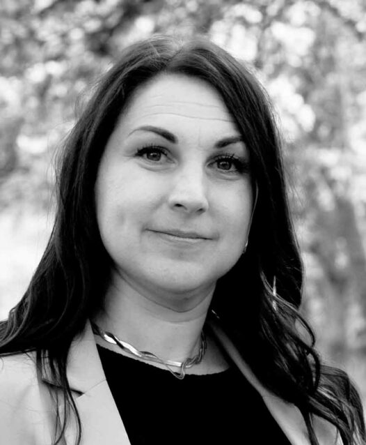 Kvinna med långt hår i kavaj ler subtilt, med oskarp bakgrund av träd.