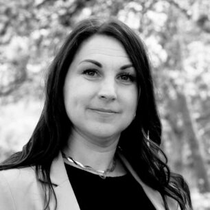 En svartvit porträttbild av en person med långt hår, iklädd kavaj, med suddig naturbakgrund.