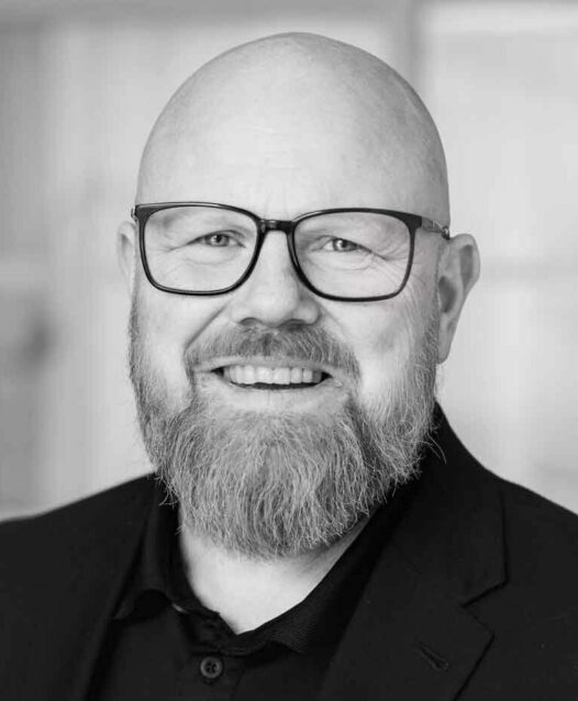En man med glasögon och skägg ler mot kameran i en svartvit porträttbild. Han bär en mörk kostym och skjorta.