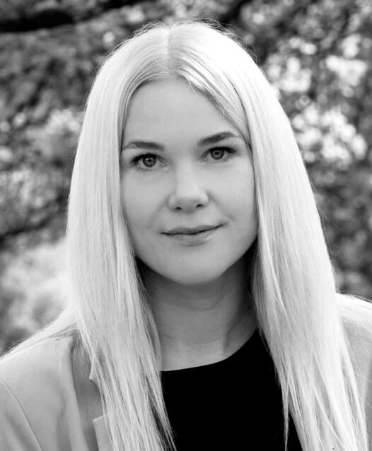 Kvinna med långt, rakt hår tittar in i kameran, med en suddig bakgrund av träd.