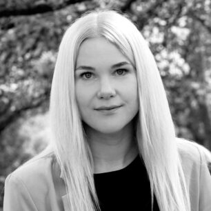 En kvinna med långt ljust hår står utomhus med suddig bakgrund av träd.