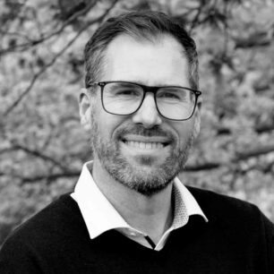 En man med glasögon och skägg ler mot kameran, med träd i bakgrunden.