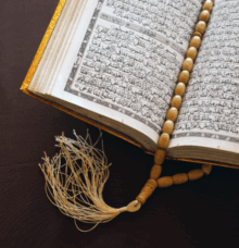En öppen bok med arabisk text ligger på en brun yta, med ett radband av träpärlor vilande på sidorna.