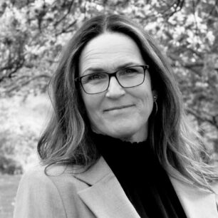 En person med glasögon ler lätt, omgiven av suddig bakgrund av träd.