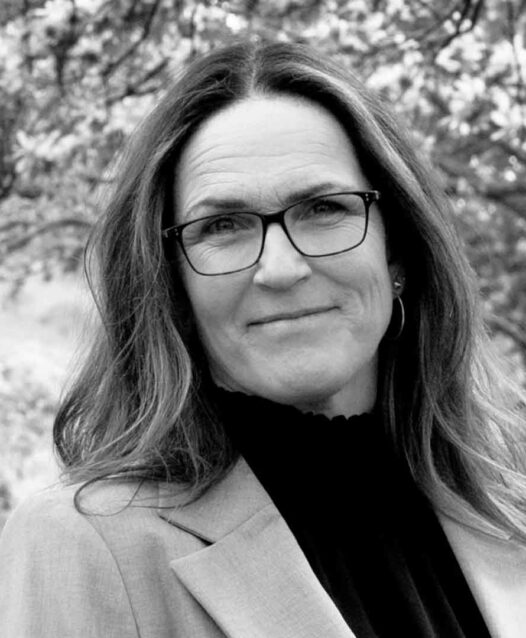 En person med glasögon ler lätt, omgiven av suddig bakgrund av träd.