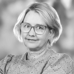 Kvinna med glasögon och kort ljust hår, iklädd en spetsmönstrad blus, tittar in i kameran med ett neutralt ansiktsuttryck. Bakgrunden är suddig.