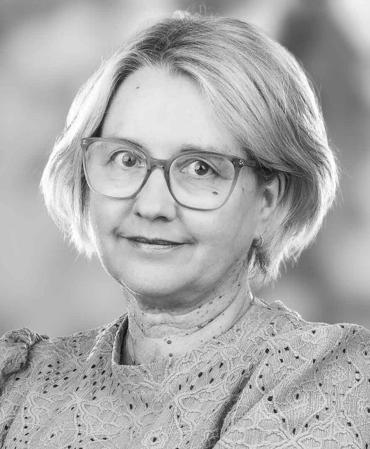 Kvinna med glasögon och kort ljust hår, iklädd en spetsmönstrad blus, tittar in i kameran med ett neutralt ansiktsuttryck. Bakgrunden är suddig.