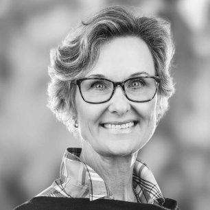 En svartvit porträttbild av en leende kvinna med glasögon och kort hår, iklädd en rutig skjorta.