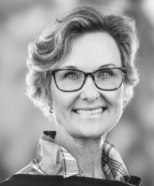 En svartvit porträttbild av en leende kvinna med glasögon och kort hår, iklädd en rutig skjorta.