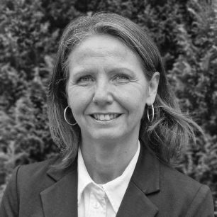 Kvinna med axellångt hår och örhängen, klädd i kavaj, ler framför en bakgrund av buskar.