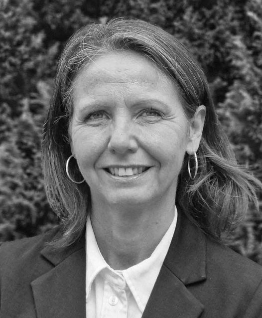 Kvinna med axellångt hår och örhängen, klädd i kavaj, ler framför en bakgrund av buskar.