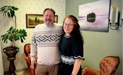 En man och en kvinna står leende i ett rum med klassisk inredning, omgivna av växter och tavlor på väggarna.