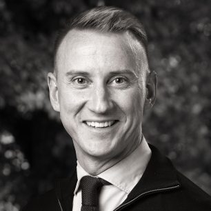 Man som ler, iklädd skjorta och slips, med suddig bakgrund av träd.