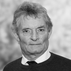 En äldre man med rufsigt hår och neutral ansiktsuttryck, klädd i skjorta och tröja, mot en suddig bakgrund.