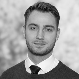 En person med kort hår och skägg, klädd i skjorta och slips, tittar mot kameran med en oskarp bakgrund.