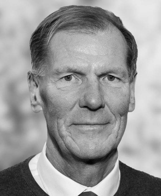 En äldre man i kavaj och slips ser in i kameran, med en neutral ansiktsuttryck och suddig bakgrund.