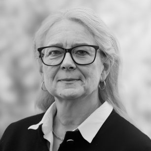 En äldre kvinna med glasögon och långt hår ser in i kameran, med en suddig bakgrund.