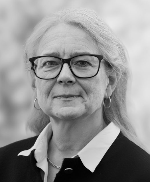 En äldre kvinna med glasögon och långt hår ser in i kameran, med en suddig bakgrund.