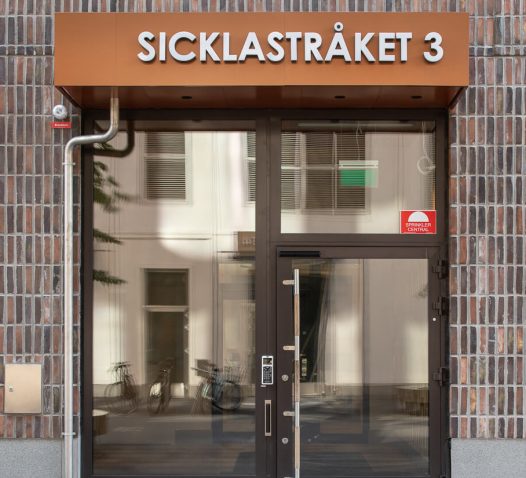 Ingång till byggnad med skylttexten "SICKLASTRÅKET 3" ovanför glasdörrarna, omgiven av tegelväggar.