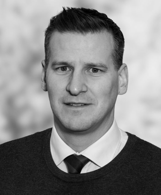 En man i kostym och slips ler svagt framför en suddig bakgrund.