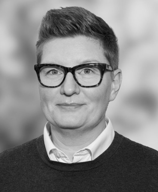 En person med glasögon och kort hår ler svagt mot kameran, klädd i en tröja och skjorta. Bakgrunden är suddig.