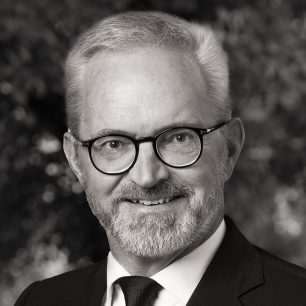 En man med glasögon och skägg ler, klädd i kostym och slips. Bakgrunden är suddig och skapar ett professionellt intryck.