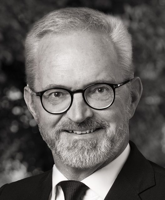 En man med glasögon och skägg ler, klädd i kostym och slips. Bakgrunden är suddig och skapar ett professionellt intryck.