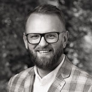 En man med glasögon och skägg ler, iklädd en rutig kavaj, mot en suddig bakgrund av träd.