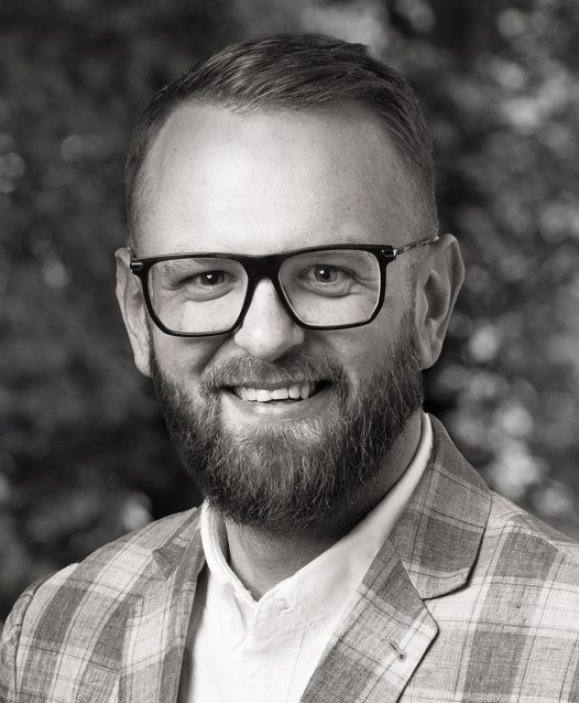 En man med glasögon och skägg ler, iklädd en rutig kavaj, mot en suddig bakgrund av träd.