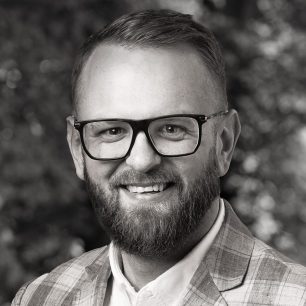 En man med skägg och glasögon ler. Han bär en rutig kavaj och en skjorta. Bakgrunden är suddig med träd och löv.