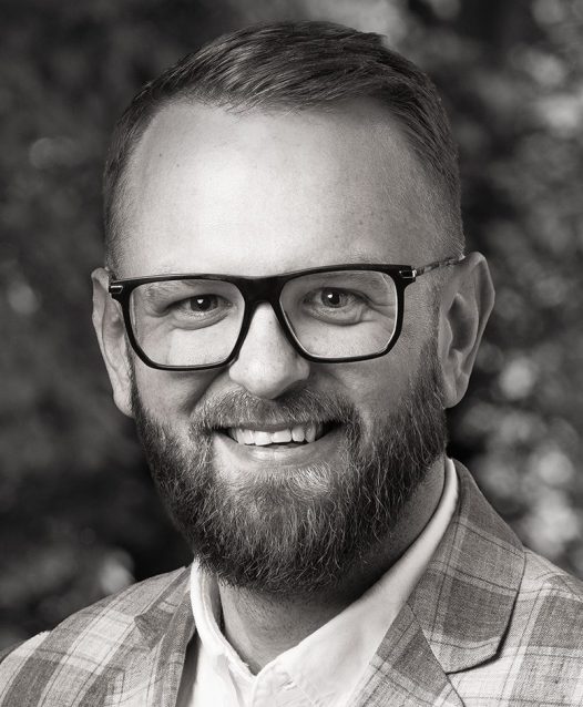 En man med skägg och glasögon ler. Han bär en rutig kavaj och en skjorta. Bakgrunden är suddig med träd och löv.