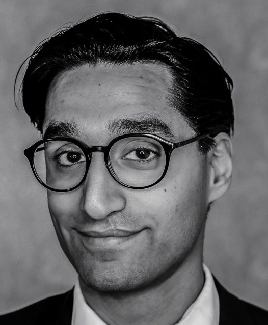 A person with short dark hair and glasses, smiling slightly in a grayscale portrait.