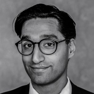 Person i svartvit porträttbild med glasögon och kostym, ler mot kameran.