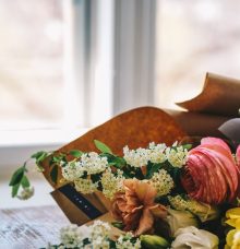 A bouquet of assorted flowers, including pink and peach roses, white blossoms, and yellow flowers, wrapped in brown paper, placed on a wooden table near a window.