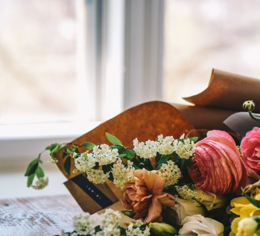 A bouquet of assorted flowers, including pink and peach roses, white blossoms, and yellow flowers, wrapped in brown paper, placed on a wooden table near a window.