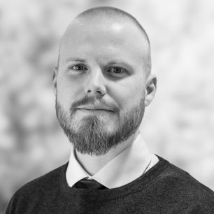 A man with a beard and shaved head wearing a sweater over a collared shirt and tie, looking at the camera against a blurred background.