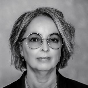 A grayscale portrait of a person with short hair and round glasses, looking directly at the camera with a neutral expression.