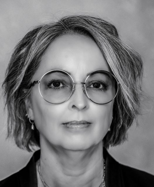 A grayscale portrait of a person with short hair and round glasses, looking directly at the camera with a neutral expression.