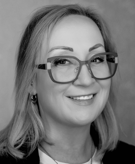 A person with glasses smiles in a black and white portrait, with shoulder-length hair and a neutral background.