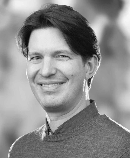 A person with short hair smiling, wearing a sweater, in a grayscale portrait.