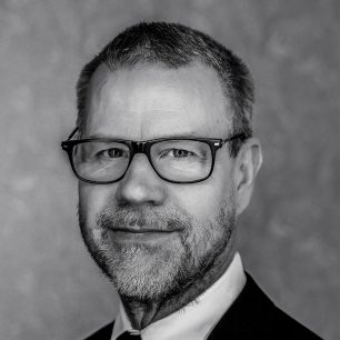 A man with glasses and a beard is wearing a suit and tie, looking directly into the camera with a neutral expression.