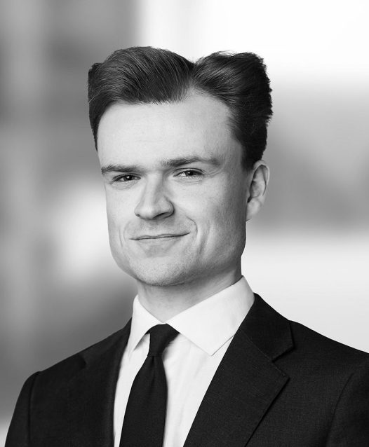 A man wearing a suit and tie smiles slightly in a black and white portrait with a blurred background.