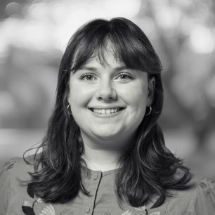 A person with shoulder-length hair smiles warmly in a black-and-white portrait, with a blurred background of trees and light.