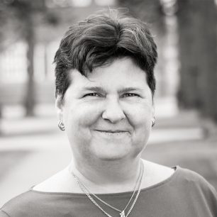 A person with short hair smiles while wearing earrings and a necklace, set against a blurred outdoor background in grayscale.