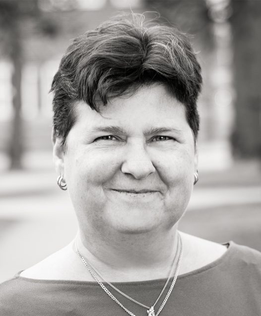 A person with short hair smiles while wearing earrings and a necklace, set against a blurred outdoor background in grayscale.