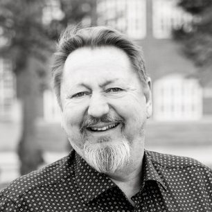 A person with a beard and mustache smiling, wearing a patterned shirt, with a blurred building in the background.