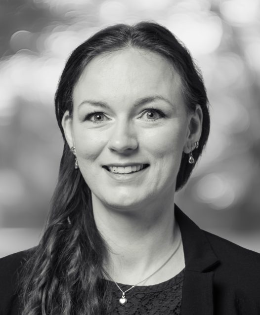 A woman with long hair, wearing a blazer and earrings, smiles in a black and white portrait with a blurred background.